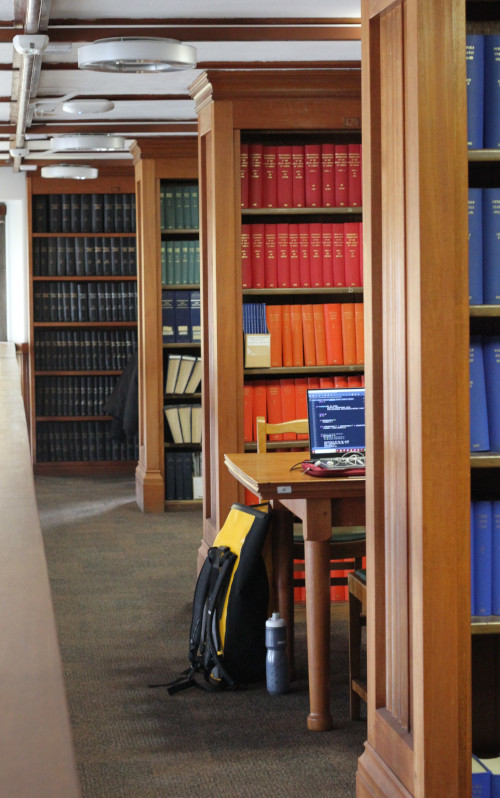 Rows of wooden bookshelves, in between the first wo is a wooden table with a laptop on it and their rucksack and water bottle next to the table on the floor. 