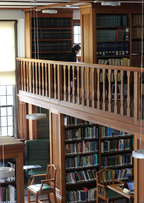 cross section of two floors of the physics library, mezzanine floor visible due to balcony banisters, both floors contain rows of bookcases with wooden desk in between, everything is lit by a window on the left. On the mezzanine floor towards the back a student is sat at a desk studying.  