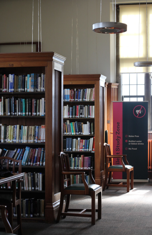 Two bookcases filled with books, both book cases have wooden chairs at the end of them, scene is lit by a window at the back right. 