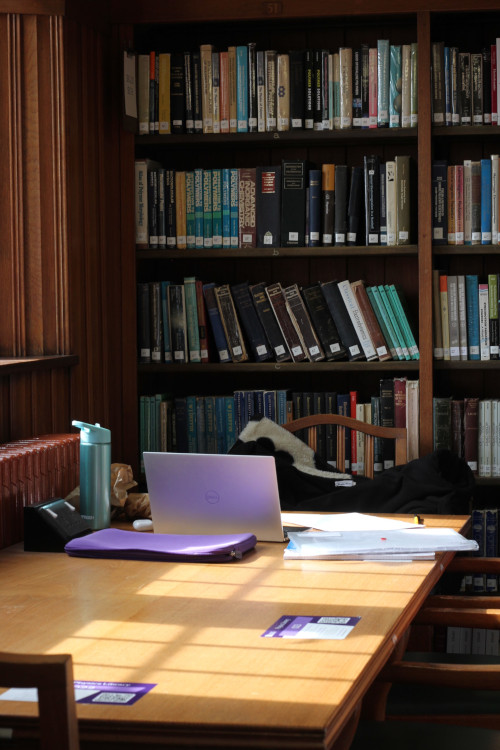 A wooden desk with an open laptop, water bottle and sheets of paper on it, behind the desk is a bookcase full of books. 