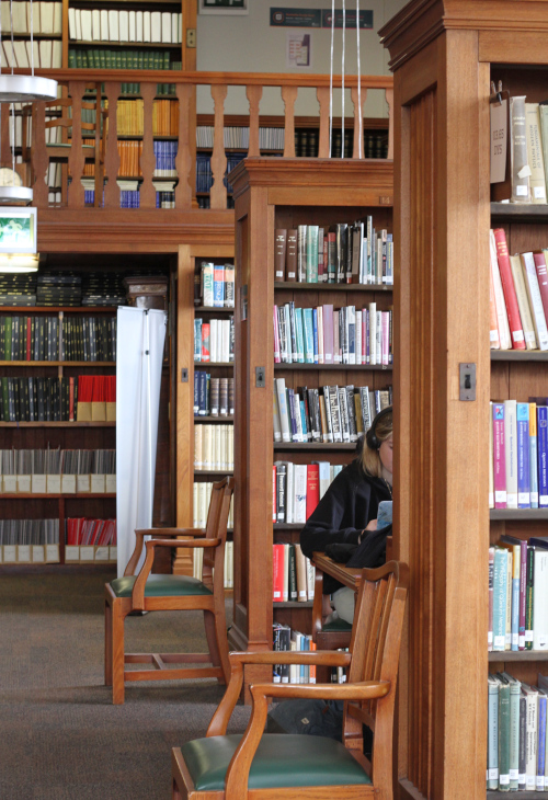 Three wooden bookcases, between the first two a student can be half seen studying at a desk, there are also wooden chairs at the end of the first two bookcases. 