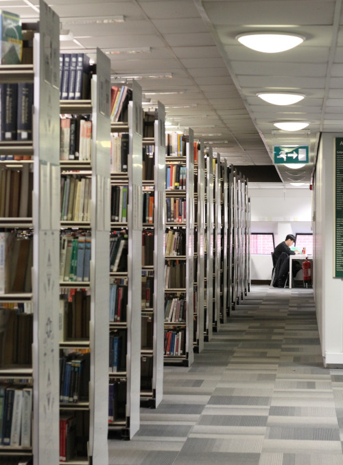 Looking down a corridor, left-hand side receding bookshelves, at the end of the corridor a student works at a desk in front of two windows. 