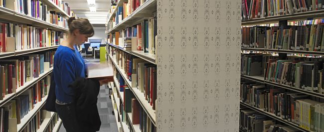 Shows the Arts and Social Sciences Library. Stacks of shelves filled with books, a person in a blue top standing between two sets of shelves reading an open book.