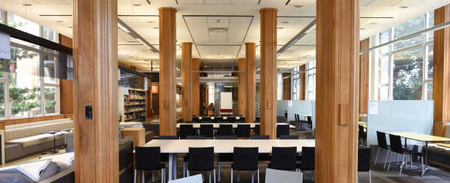 Shows the Medical Library. Large long and narrow open plan room filled with wooden columns and large group desks with chairs, with windows on the side walls.