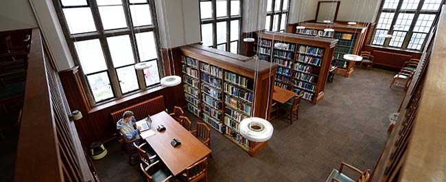 Birds eye view of the Physics Library, a wood panelled library with shelving with books, desks and one person studying with a laptop.