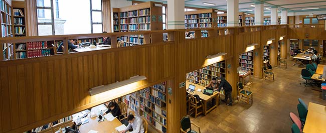 Shows the Queens Library. Two story library with wooden shelves making alcoves for desks, lots of students working at desks.