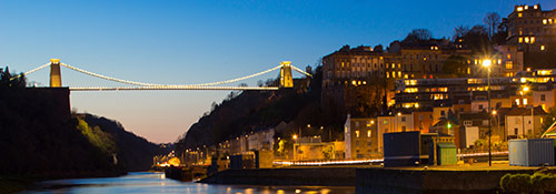 The iconic Clifton Suspension Bridge over the Avon Gorge at night. The bridge and nearby houses are lit up.