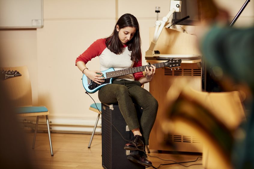 A female student with long hair in a red and white top sat of a black box, looking down and strumming a small guitar.  