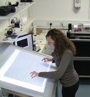 The multi-touch table being used to move particles that are located under the microscope on top of the Dynamic Holographic Assembler, the black machine in the background.