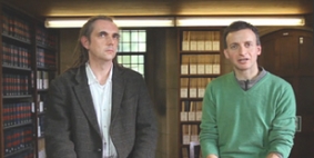 Two white men stood together in a room with bookcases behind them.