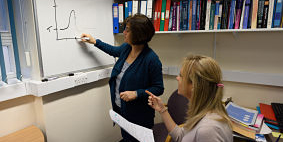 Woman in a suite jacket drawing a curved line graph on a whiteboard in an office, while another woman sits nearby pointing at the graph and holding a paper. There are ring binders and books on a shelf on the wall and on the desk behind the seated woman. 