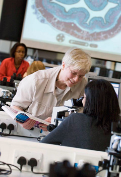 A tutor showing a student something in a textbook. The student is using a microscope in a lab, where there are several rows of microscopes and other students also working. 