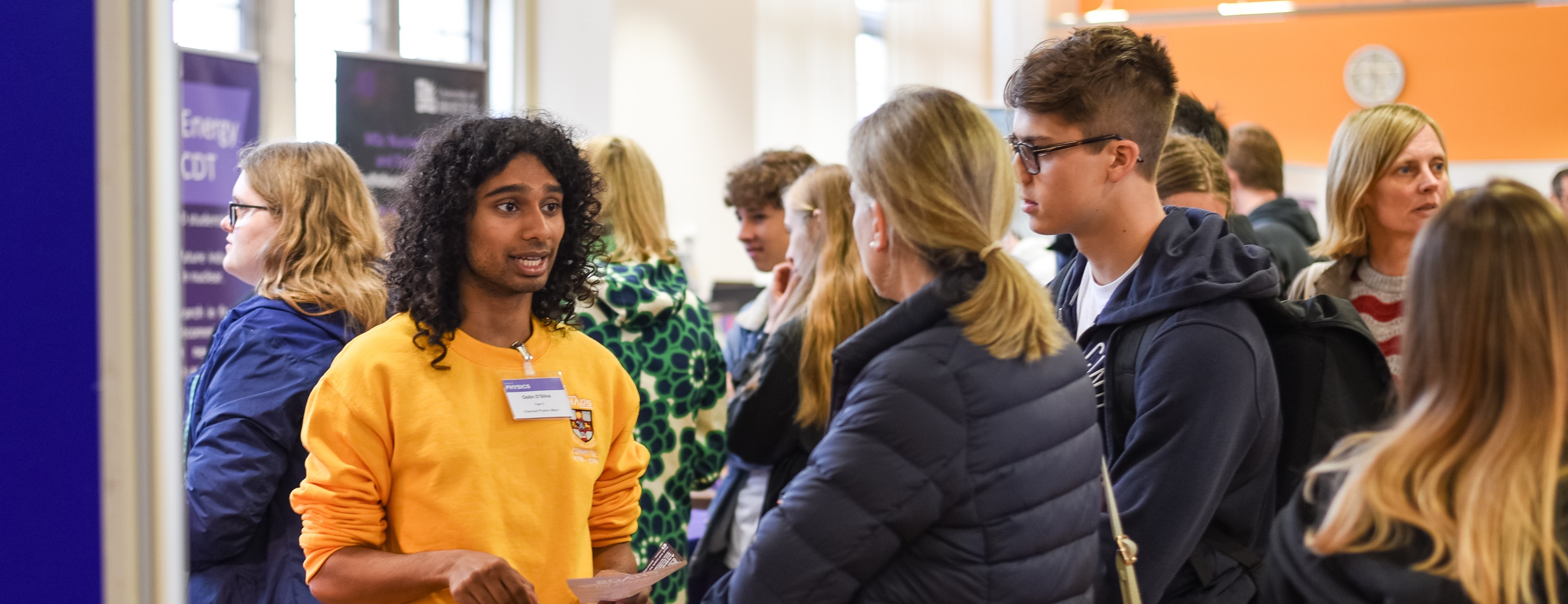Student talks to prospective student at an Open Day event. In the background there are other prospective students and their parents walking about.