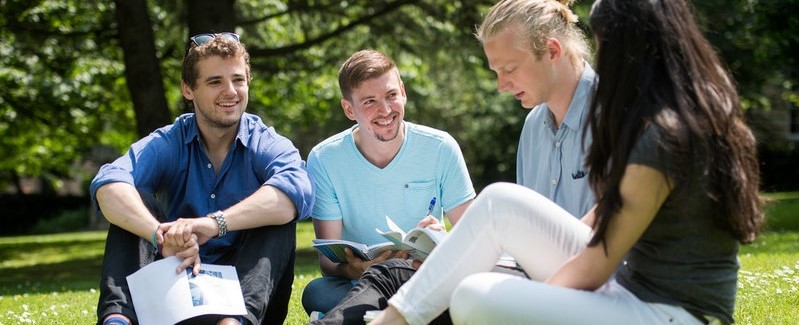 Four people in conversation sit on grass. Three of them are holding papers. 