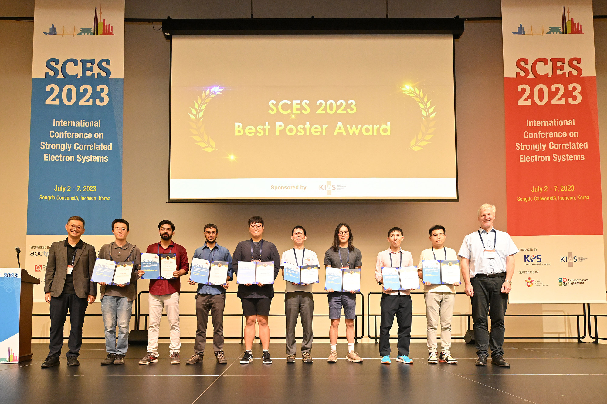 Entrants of the SCES 2023 Poster Prize stand in a row holding certificates. Behind them is a projection which says 'SCES 2023 Best Poster Prize', which is flanked either side by large SCES 2023 banners.
