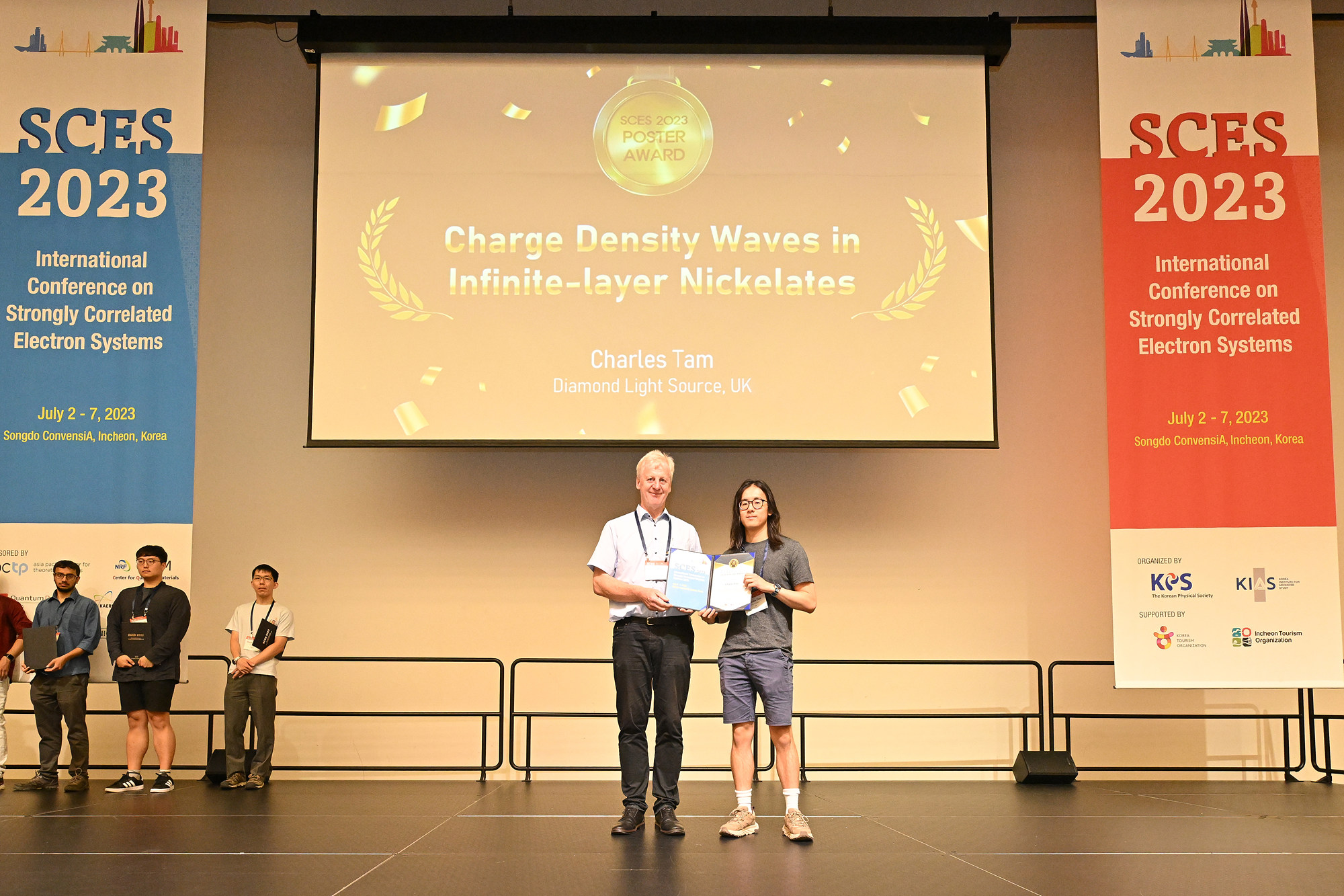 Charles Tam accepts his SCES 2023 poster prize. He is stood on stage next to another person; both are holding the certificate. The projection behind them reads 'Charge Density Waves in Infinite-layer Nickelates.' This is flanked by large SCES 2023 banners on both sides.