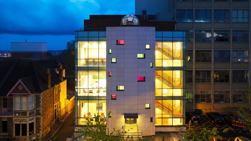 The NSQI centre is lit up at dusk. Orange light shines through the windows. A section of the HH Wills Physics Building is visible in the right of the frame.