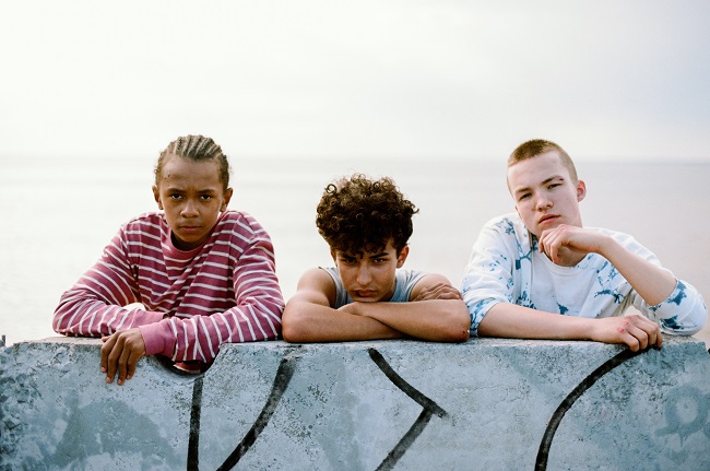 Three teen boys resting their arms on a graffiti-marked wall and staring at the camera.