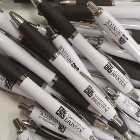 Close up image showing a pile of University of Bristol branded Contour Eco pens, with the logo printed in black onto a white barrel with a matching black grip