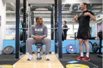 A male personal trainer talking to an adult male who is sat down on a bench within a squat rack, in the gym.
