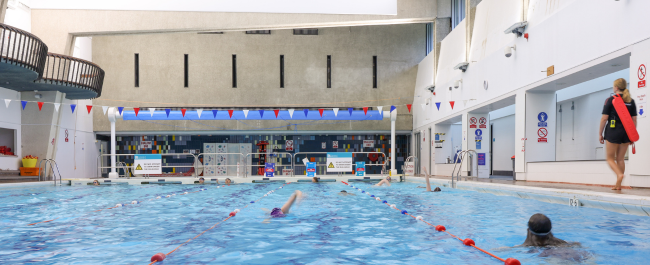 Swimmers in various lanes at the University of Bristol swimming pool during a lesson as a lifeguard walks along the side of the pool