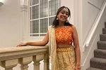 Current student Manisha smiling, wearing a bright orange and gold sari standing in front of a stone staircase and large window. 