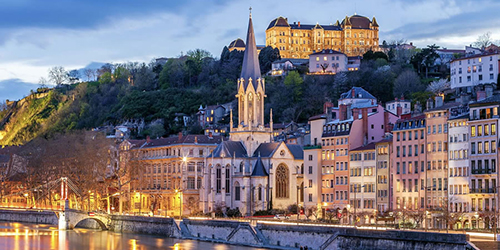 A church and surrounding buildings by a river in a European city are lit up at night.