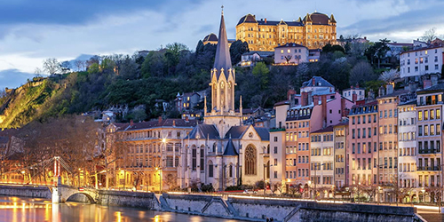A church and surrounding buildings by a river in a European city are lit up at night.