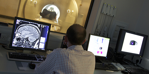 A person looking at a computer showing a scan of someone's brain, with an MRI machine in the background.