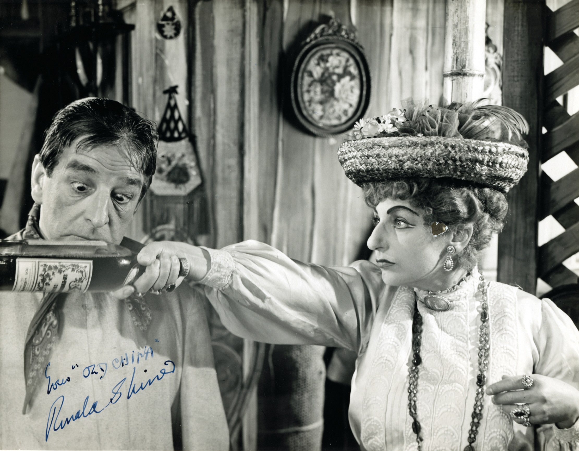 Joseph looking at a bottle held by Madame Parole in her right hand with arm stretched out. Photograph signed by Ronald Shine.