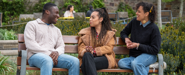 Three people of different genders sitting on a bench in a garden, smiling and talking to each other.