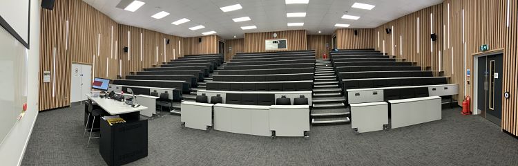 A spacious and modern lecture theatre with tiered seating. A top table and projector screen setup at the front of room. 