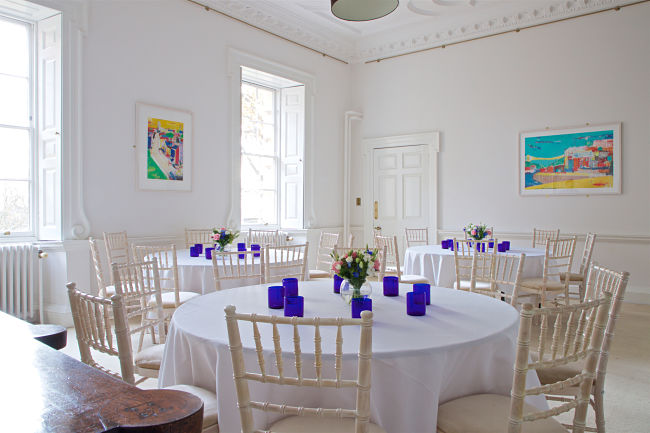 A bright room with white walls and woodwork and period feature. Setup with round tables for dinner and floral centerpieces. 