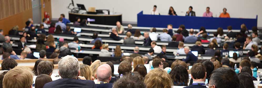 Delegates sit in a lecture theatre with tiered seating while a panel of 5 speakers present. 