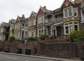 A row of terraced two-storey red brick houses. There are steps with bannisters leading up to the house entrances from the pavement, and recycling bins on the pavement outside.
