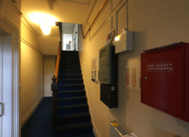 A stairwell with a bannister and 15 steps inside a house. There are several metal boxes on the wall at the bottom of the stairs, including one with 'fire safety documents' written on it.