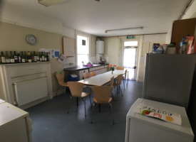 A kitchen with a washing machine and fridge freezer. There are two tables in the centre of the room with nine chairs around them.