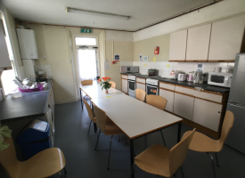 A kitchen with two ovens and hobs, a washing machine, a fridge freeze and lots of cupboards. There are two tables in the centre of the room with nine chairs around them.