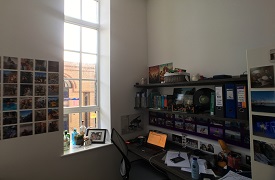 A desk with an office chair next to it and a laptop on it, plugged in at the wall. There are two shelves above the desk and lots of photos on the walls.