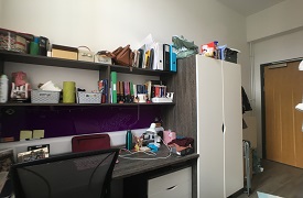 A desk with an office chair next to it. There are two shelves above the desk and a clothes cupboard next to it.