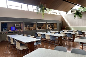 A dining hall with tables and chairs around each of them and a food serving area.