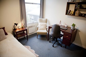 A room with a single bed in one corner and an armchair and desk opposite in a catered room.