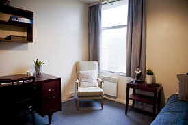 A room with a single bed in one corner and an armchair and desk opposite in a catered room.