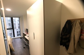 A room with a coat rack and wardrobe against the wall. Beyond it there is a bed and a desk with a chair in the main room.