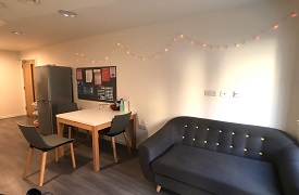 A room with a fridge freezer, a table, and a sofa against one wall. The table has three chairs around the other sides.