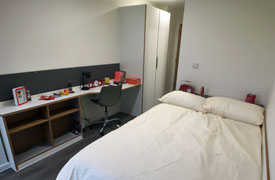 A room with a double bed in one corner and a desk with an office chair against the opposite wall. The desk also has a set of shelves beneath it and a wardrobe next to it.