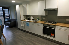 A kitchen with an oven and hob, a sink, several cupboards and drawers, and a fridge freezer. There is also a sofa next to the fridge at the far end of the room.