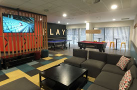 A room with a semi-circular sofa facing a coffee table and a wall-mounted television. At the back of the room there is a pool table, and a ping pong table next to a wall with the word 'play' written on it.