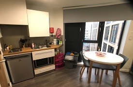 A room with an oven, fridge, sink and some cupboards against one wall. There is a table nearby with two chairs around it.