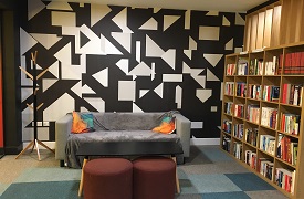 A room with a large bookcase against one wall, and a sofa and coatstand against the adjacent wall. There is a table and two small stools in front of the sofa.